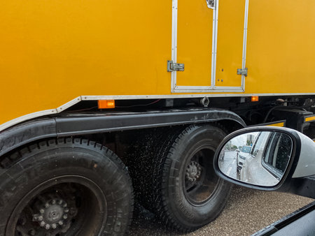 This view showcases the impressive size of a large yellow trucks heavyduty tires next to a cars side mirror, capturing the industrial aesthetics typical of highways and roadwaysの写真素材