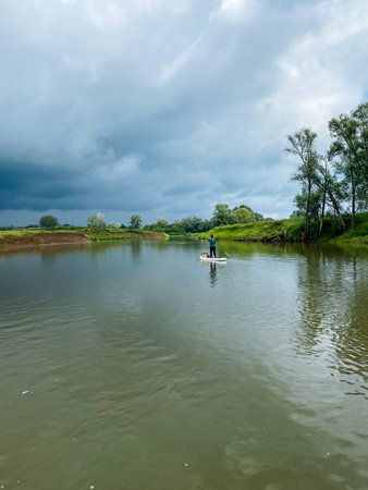A confident individual skillfully balances on a paddle board, gliding over a peaceful river while appreciating the stunning beauty of nature, including the vibrant sky and lush greeneryの写真素材