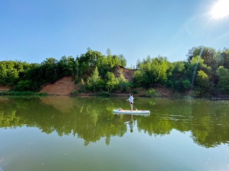 In the center of a serene lake, a man is comfortably standing upright on a paddle board, enjoying the peaceful surroundings and engaging in a popular recreational activity on the waterの写真素材