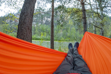 A person lounges in a vibrant orange hammock between two trees, with a tranquil river flowing in the background, capturing the essence of outdoor relaxation and escape from daily lifeの写真素材