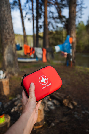 In a serene camping setting, a hand grips a red first aid kit, showcasing the need to be prepared...
