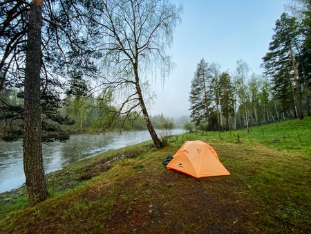 A vibrant orange tent is comfortably situated on a lush grassy hill that overlooks a stunningly beautiful river, creating a picturesque and serene scene in nature that is truly breathtakingの写真素材