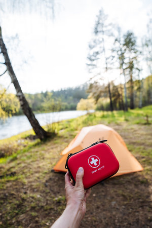 A hand holding a red first aid kit in the foreground, with an orange tent by a tranquil lake surrounded by lush trees. Perfect for outdoor enthusiasts and safety-conscious campers.の写真素材
