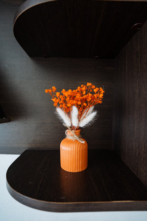 On a shelf in the room, there sits a small orange vase that is beautifully filled with an assortment of dried flowers, creating a charming decorative piece that brightens the spaceの写真素材
