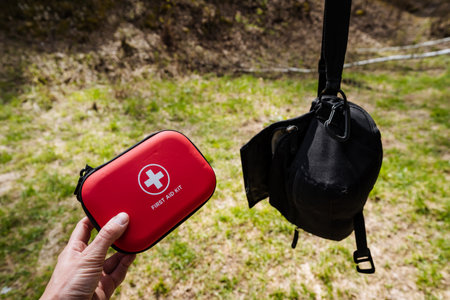 A hand firmly grasping a vibrant red first aid kit next placed to a sturdy black tactical bag, making it an ideal setup for outdoor adventures, hiking excursions, and thorough emergency preparednessの写真素材