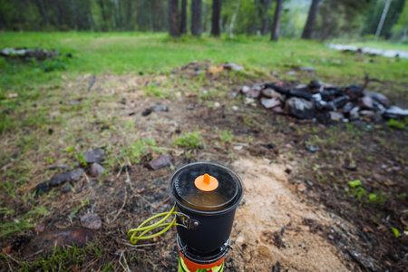 A portable camping stove sits on the grass, surrounded by trees, with a cozy fire pit smoldering nearby. This scene delights outdoor cooking enthusiasts who appreciate natures beautyの写真素材