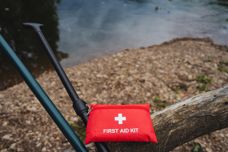 A bright red first aid kit sits on a log by the calm water, highlighting the importance of safety during outdoor activities like fishing, camping, or hiking where preparedness is essentialの写真素材