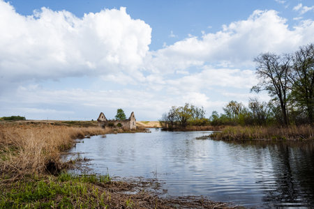 A serene riverside scene features weathered ruins among vibrant grasslands and fluffy clouds under a clear blue sky, creating a peaceful ambiance ideal for relaxation and natures beautyの写真素材