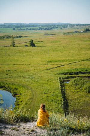A woman wearing a bright yellow dress is gracefully sitting atop a beautiful hill while enjoying the view of a vast green field that stretches out before her, creating a picturesque sceneの写真素材