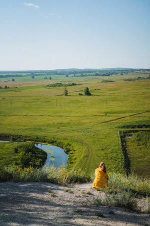 A woman wearing a vibrant yellow dress is seated on top of a grassy hill, enjoying the view of a winding river flowing below. The landscape is picturesque, filled with natural beauty and serenityの写真素材