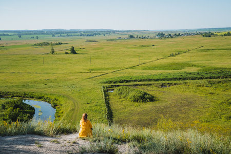 A woman wearing a bright yellow dress is comfortably seated on top of a scenic hill, enjoying the view of a vast grassy field that stretches out before her, bringing peace to her surroundingsの写真素材