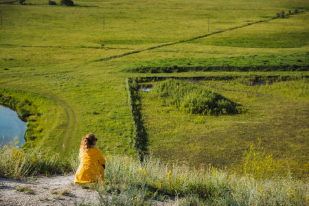 A woman wearing a bright yellow jacket is comfortably seated atop a scenic hill, where she enjoys the beautiful view of a winding river below her, surrounded by lush greenery and natureの写真素材
