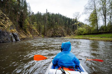 A tranquil scene of a person kayaking on a calm river, surrounded by lush trees and rugged cliffs,...