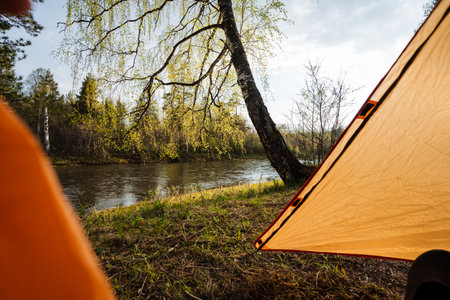 Experience a peaceful camping trip by the riverside, surrounded by lush greenery and a bright orange tent under a clear blue skyideal for nature lovers and outdoor enthusiasts seeking adventureの写真素材