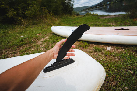 A person holds a sleek black fin, preparing to install it on a white paddleboard by a tranquil lake, surrounded by lush greenery, ideal for themes of outdoor adventure and explorationの写真素材