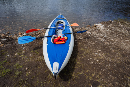 A bright blue kayak rests on the sandy shore by a calm river, accompanied by vibrant paddles and essential safety gear, beckoning adventurers to embark on an exciting day filled with fun on the waterの写真素材