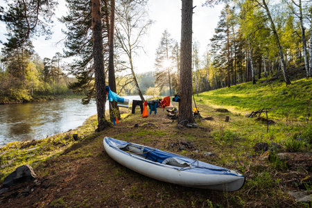 A tranquil scene of a kayak on a serene river shore, surrounded by lush greenery and colorful clothes hanging to dry, ideal for camping and outdoor adventures with friends and familyの写真素材