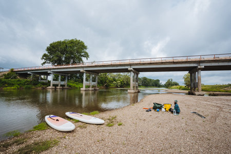 In a serene and peaceful river scene, paddleboards rest idly on the sandy shore, positioned comfortably beneath a sturdy bridge, surrounded by vibrant lush greenery and softly overcast skies aboveの写真素材