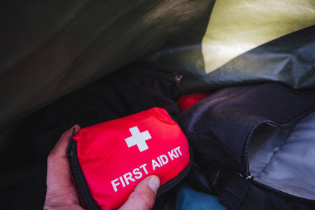 A closeup of a hand holding a vibrant red first aid kit amidst camping gear, promoting safety and preparedness for outdoor adventures and emergenciesの写真素材