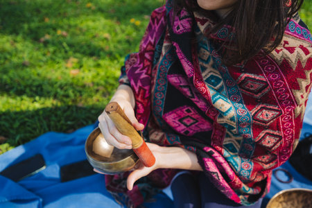 In a serene and tranquil moment captured beautifully in nature, a woman wrapped in a vibrant and colorful shawl gently plays a singing bowl, which promotes relaxation and mindfulness effectivelyの写真素材