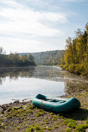 A vivid green inflatable boat rests on a sandy shore by a tranquil river, surrounded by lush autumn foliage, creating an idyllic spot for fishing, hiking, or simply enjoying natureの写真素材