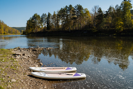 Two colorful paddleboards lean against a rocky shoreline by a tranquil river, surrounded by stunning autumn foliage reflecting on the clear water under a blue sky, perfect for outdoor activitiesの写真素材