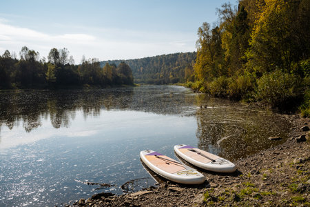 In a tranquil autumn landscape, two paddleboards rest on the riverbank, surrounded by colorful foliage, with waters reflecting beauty, inviting outdoor adventures and exploration in natureの写真素材