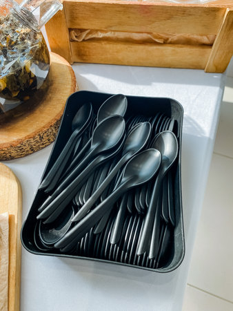 A neatly arranged tray filled with black plastic spoons and forks is placed on a wooden table, ready to be used for serving meals, making it perfect for any dining occasion or eventの写真素材