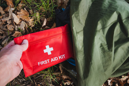 A hand firmly grips a vibrant red first aid kit nestled amidst a backdrop of lush, thriving nature, symbolizing the vital importance of safety and emergency preparedness for all outdoor adventurersの写真素材