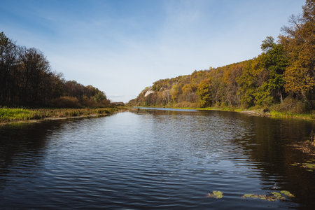 A peaceful river flows under a clear sky, surrounded by vibrant autumn leaves on rolling hills, creating a picturesque scene perfect for relaxation and an escape into natures beautyの写真素材
