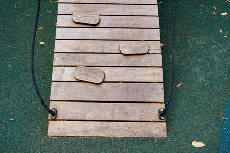 A strong and sturdy wooden plank bridge that is constructed with natural stones, specifically designed for children to play on safely, set against a vibrant green safety mat at a colorful playgroundの写真素材