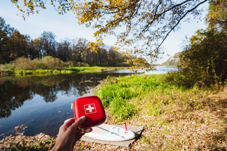 A vibrant scene shows a person with a first aid kit by a tranquil river, surrounded by colorful autumn foliage and a paddleboard, ideal for outdoor lovers seeking adventure in natureの写真素材