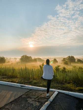 A woman stands elegantly by a quiet road, lost in thought as she gazes at a breathtaking sunset that paints the sky with vibrant colors, filling the scene with wonder and tranquilityの写真素材