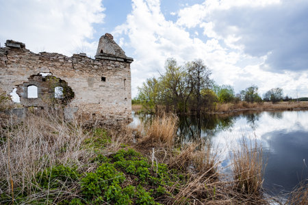 In this captivating scene, an old and crumbling building stands next to a serene pond, embraced by vibrant lush grass and majestic trees under a softly cloudy sky, enhancing the tranquil atmosphereの写真素材