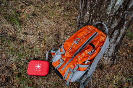 A bright orange hiking backpack leans against a sturdy tree in the forest, with a compact red first...