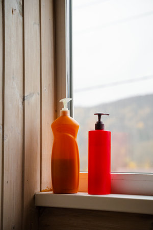 Two vibrant liquid soap bottles, one orange and one red, resting on a windowsill. The blurred outdoor scenery offers a serene background, perfect for wellness or home decor concepts.の写真素材