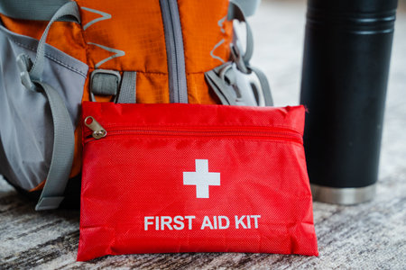 A compact red first aid kit with a white cross beside a rugged backpack and sturdy thermos is ideal...