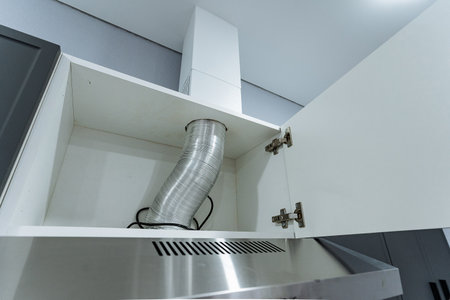 metal flex duct above range, closeup of vent connection inside cabinet, stainless hood edge and cabinet hinge visible, maintenance and installation detail for kitchen exhaust systemの写真素材