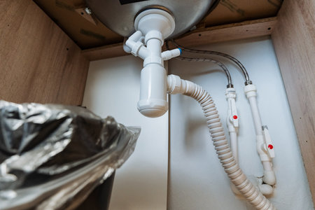 This is a detailed view of the undersink plumbing arrangement, showcasing a waste disposal unit, various water supply lines, and an efficient drainage system in a modern kitchen environmentの写真素材