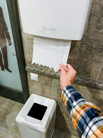 A person is in the act of pulling a roll of toilet paper out of a paper towel dispenser that is designed for such purposes, showing a common interaction with paper products in a restroom settingの写真素材