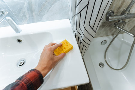 hand wiping white countertop next to bathtub with yellow sponge, shower fittings and mosaic tile visible, careful finishing of renovation, tools and materials suggested nearby, methodicalの写真素材