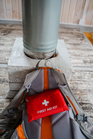 A bright red first aid kit, labeled FIRST AID, is placed in an open outdoor backpack near a metal heating unit, highlighting the importance of safety and preparedness in the wildernessの写真素材