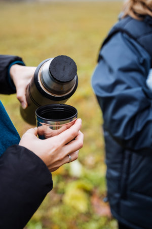 hikers pour and share steaming hot drinks outdoors, outdoor hikers pouring hot drinks into cups amidst grassy scenery, group of hikers sharing hot drinks from thermos with steam risingの写真素材