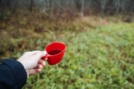 white hand extends red cup across meadow, warm drink shared as white hand offers red cup across meadow, white hand graciously extends red cup filled with warm beverage across meadowの写真素材