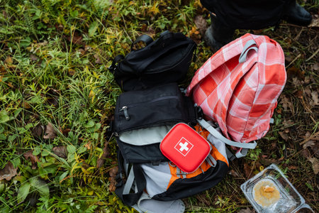 emergency outdoor supplies, first aid kit near backpack, red first aid kit beside snack and backpack on grass, emergency supplies including first aid kit and snacks on outdoor grassy areaの写真素材