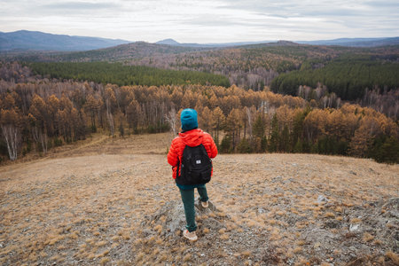 Traveler surveys scenic landscape, Explorer views peaceful outdoor surroundings, Hiker observes tranquil nature from high ridge, Adventurer gazes over lush valley with vibrant foliageの写真素材