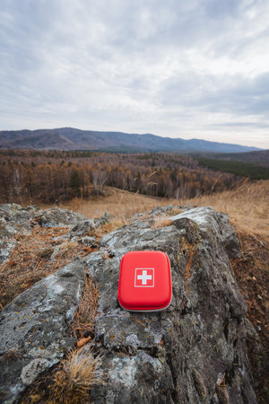 Vertical red first aid kit on summit rock with wide valley view, bright compact case framed against open sky, clouds above distant peaks, sense of preparedness for remote trekkingの写真素材