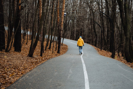 Lonely walker in yellow coat progressing down curved wet road, distant silhouette shrinking into tree line, brisk stride, hands in pockets, autumn chill evident, emotive scene of transitionの写真素材