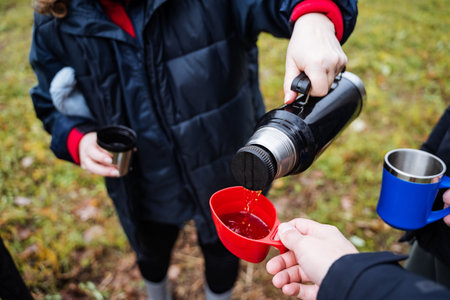 hikers in navy jackets sharing warm beverage outdoors on grassy trail near red and blue mugs