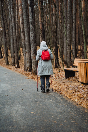 Caucasian senior woman with red backpack using walking poles on paved forest path, gray coat and hood, determined posture, autumn leaves and wooden benches nearby, resilient active agingの写真素材